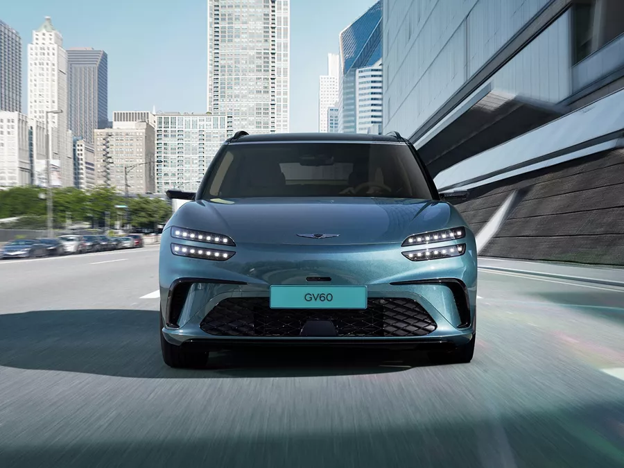 2026 Genesis GV60 in Tomso Green driving toward viewer on city road with skyline in background