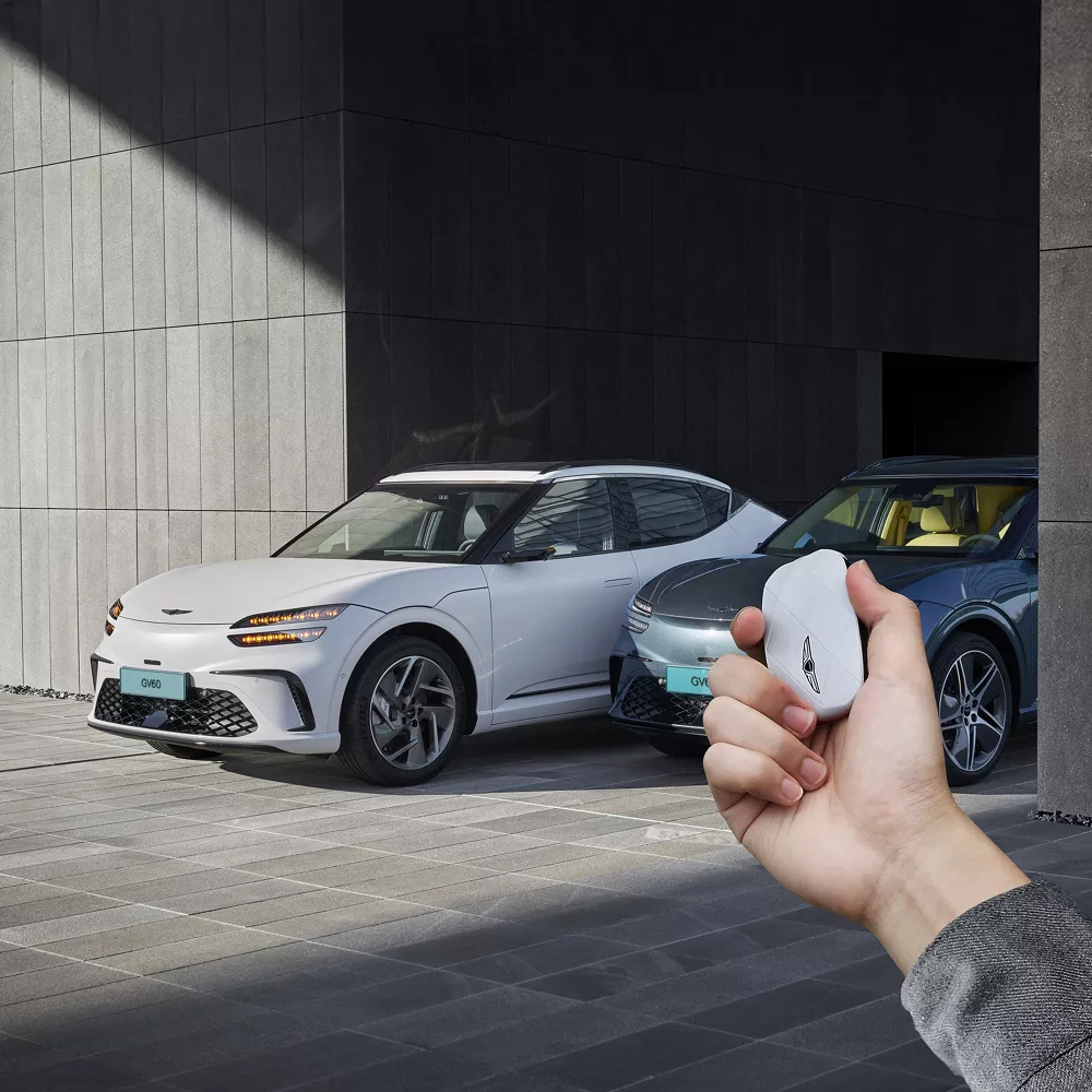 Two 2026 Genesis GV60's parked in modern garage, hand holding white Genesis key fob in foreground