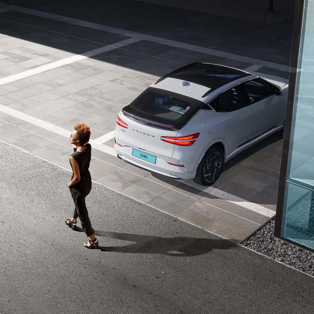 Rear view of Genesis GV60 parked as woman walks behind, highlighting rear collision avoidance feature