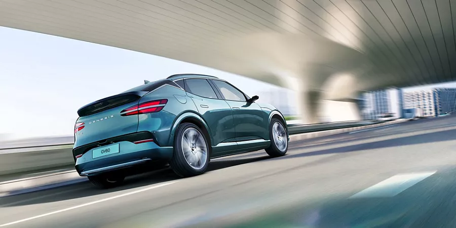 3/4 rear passenger side view of 2026 Genesis GV60 in Tromso Green driving under modern bridge