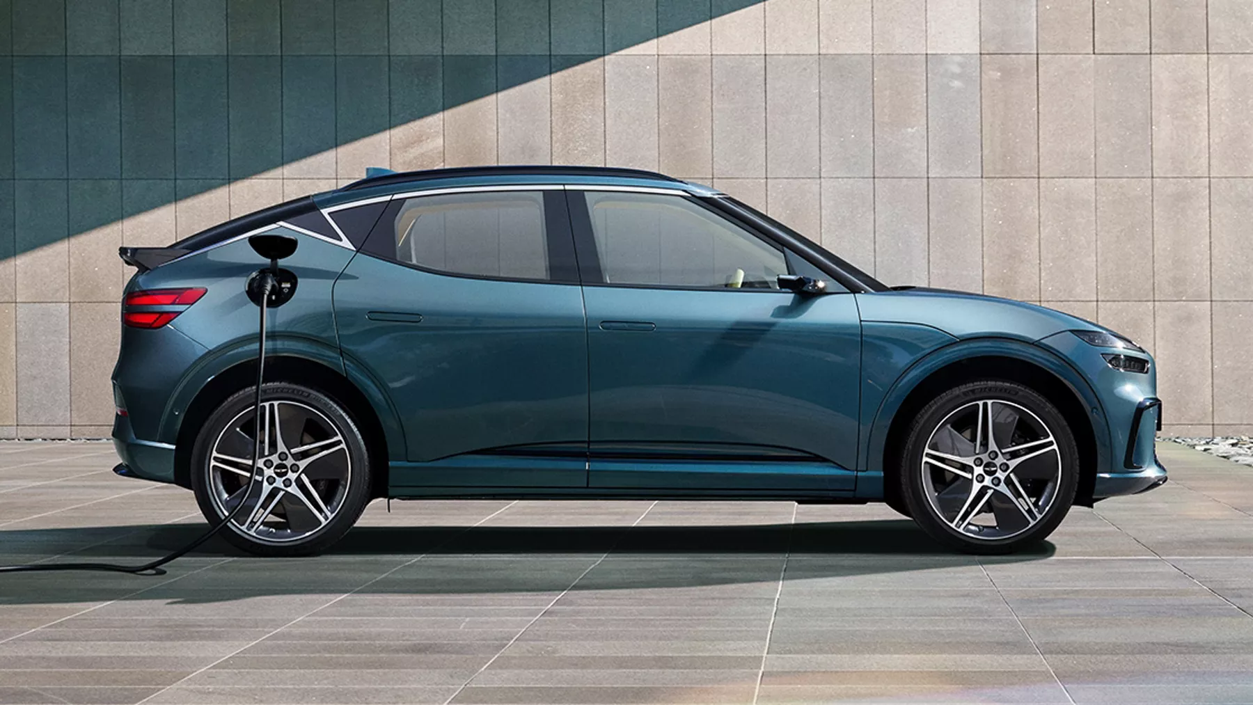 Passenger side profile view of 2026 Genesis GV60 in Tromso Green charging beside modern building
