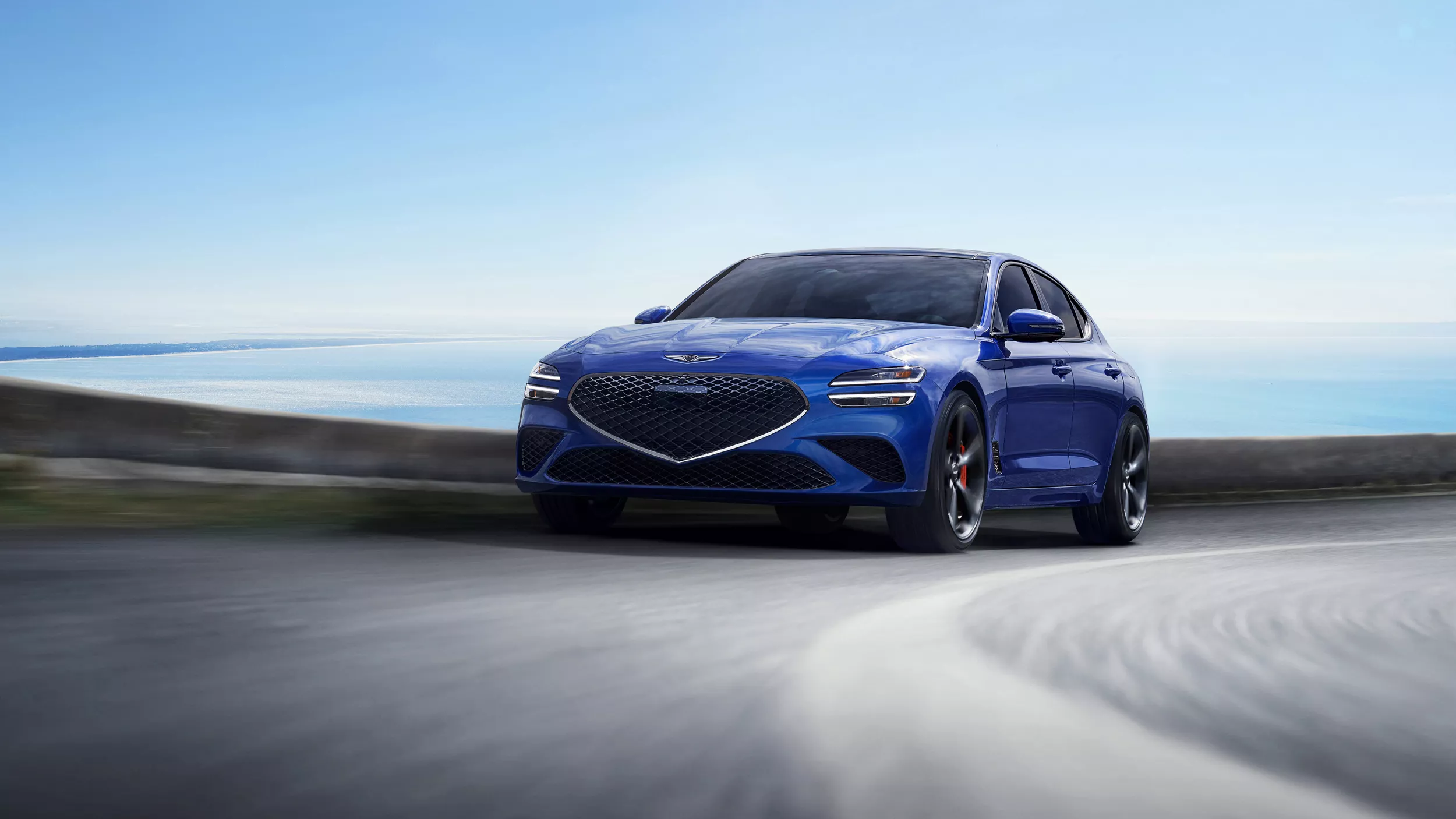 Front driver side action shot of Genesis G70 in Kawah Blue driving on a coastal highway