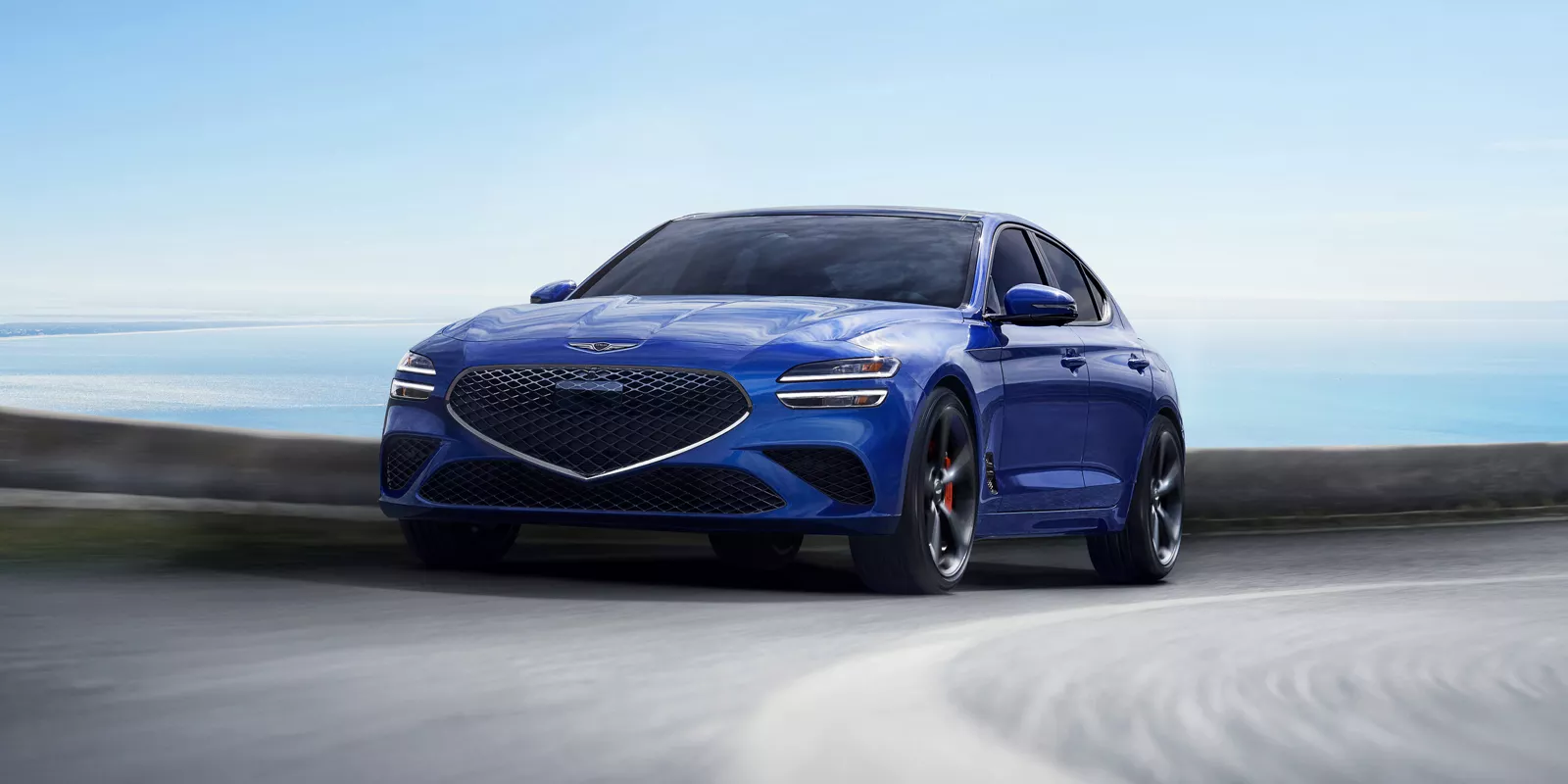 Front driver side action shot of Genesis G70 in Kawah Blue driving on a coastal highway
