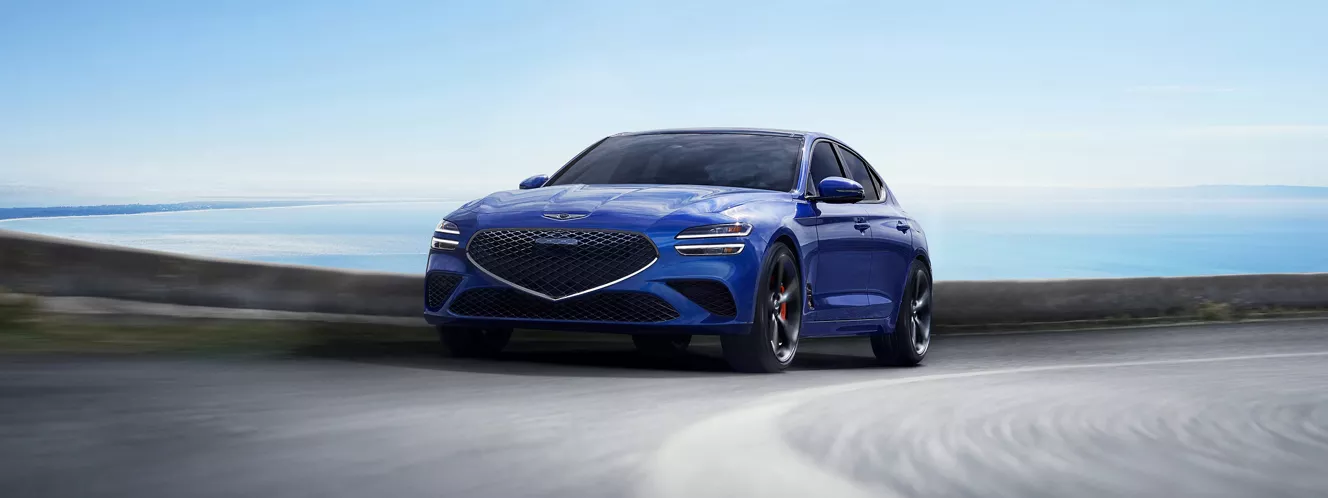 Front driver side action shot of Genesis G70 in Kawah Blue driving on a coastal highway