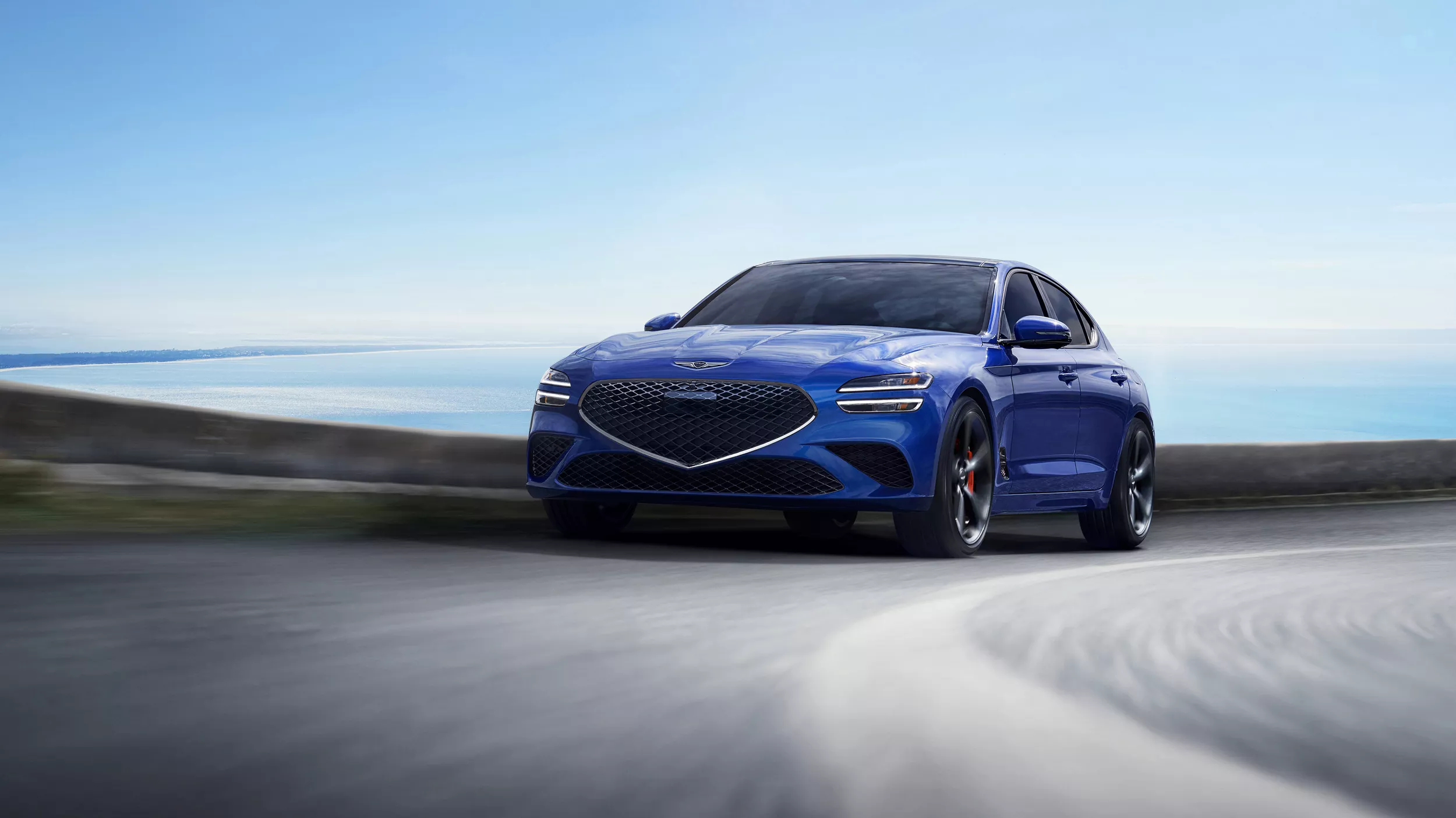 Front view of blue 2026 Genesis G70 rounding a corner with the ocean and sky in the background