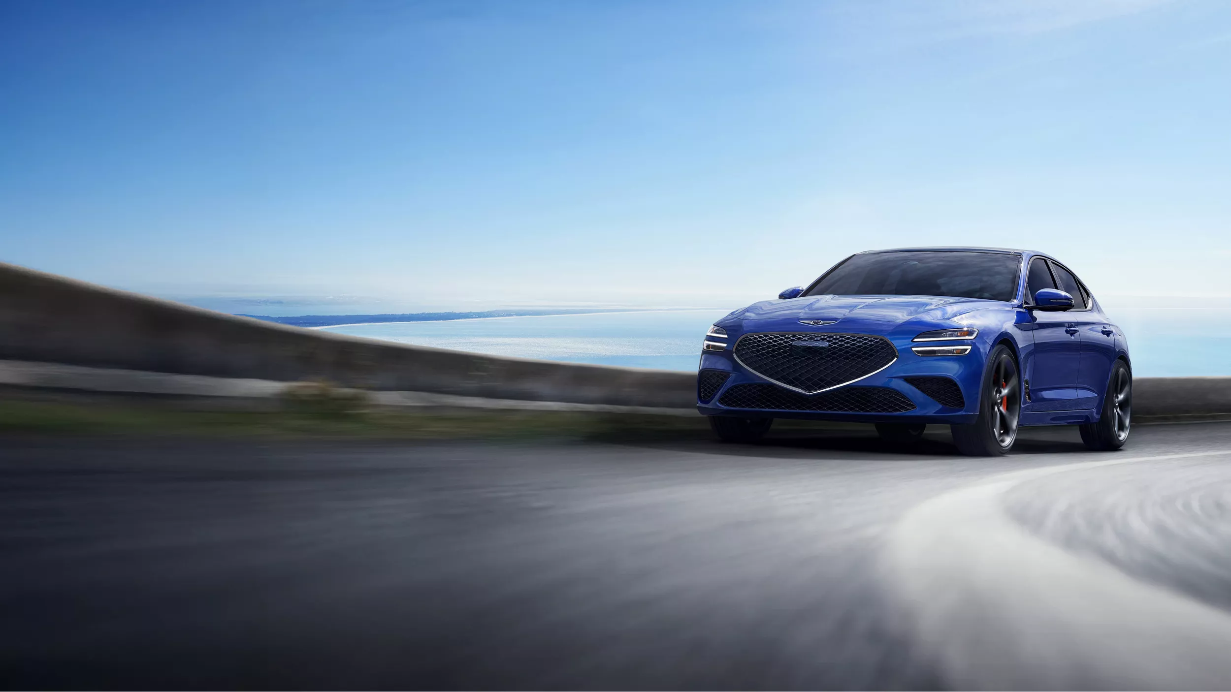 Front view of blue 2025 Genesis G70 rounding a corner with the ocean and sky in the background