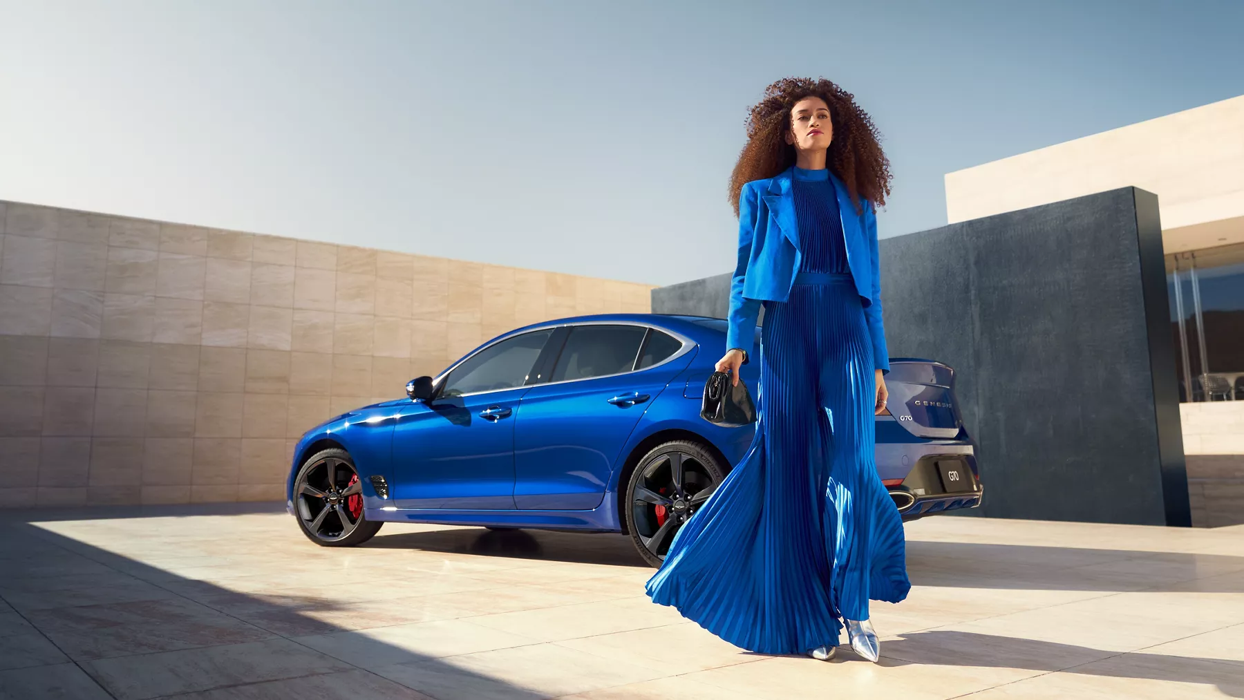Driver side view Genesis G70 in Kawah Blue parked on rooftop with woman walking in forefront