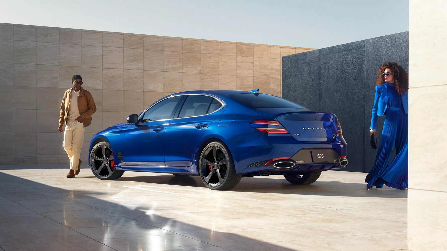 Rear driver side view of Genesis G70 in Kawah Blue as man in brown and woman in blue stand close by
