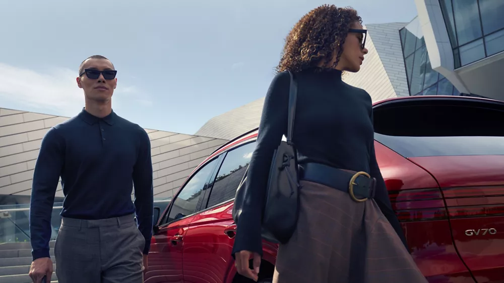 Rear view of Genesis GV70 in Mauna Red, parked in front of modern building, man and woman walk past