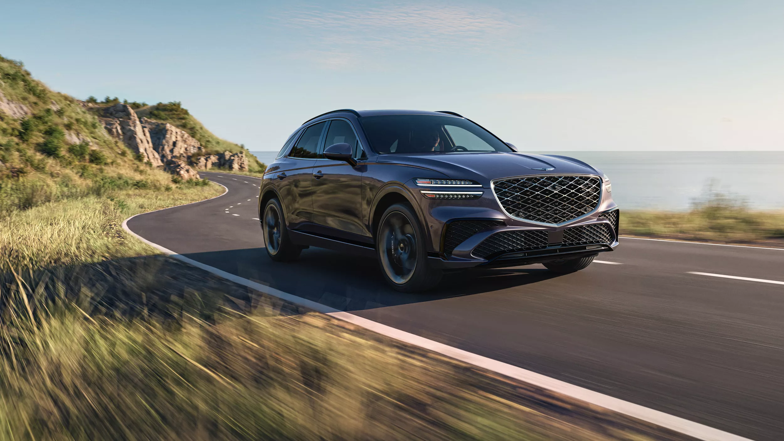 Front passenger side view of a 2026 Genesis GV70 driving along a mountainous coastal highway