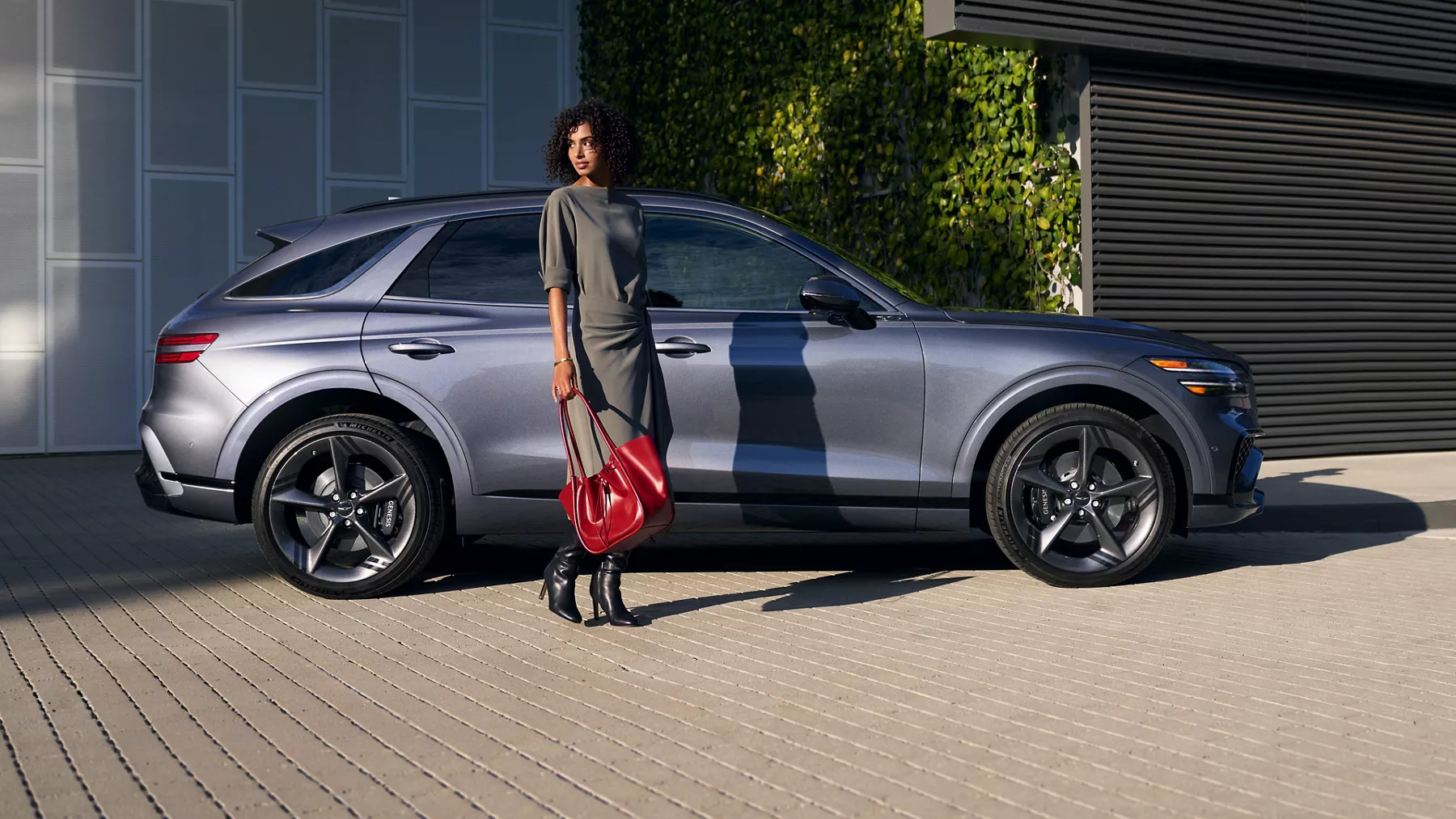 Passenger side profile of woman standing next to 2026 Genesis GV70 in front of contemporary building