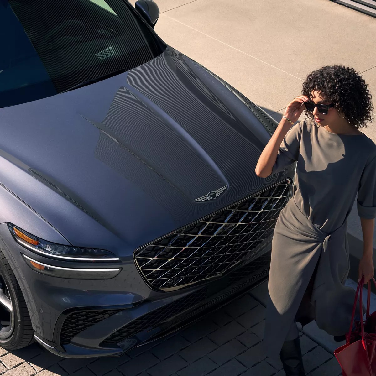 Aerial view of 2026 Genesis GV70 in gray, passenger door open, woman walking in front of vehicle