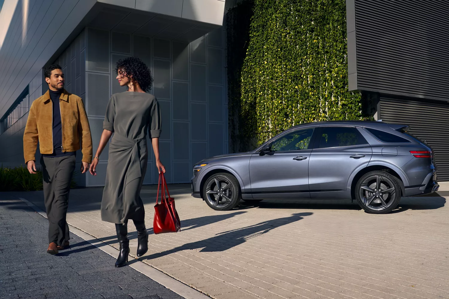 Driver side profile of couple standing next to 2026 Genesis GV70 in front of contemporary building