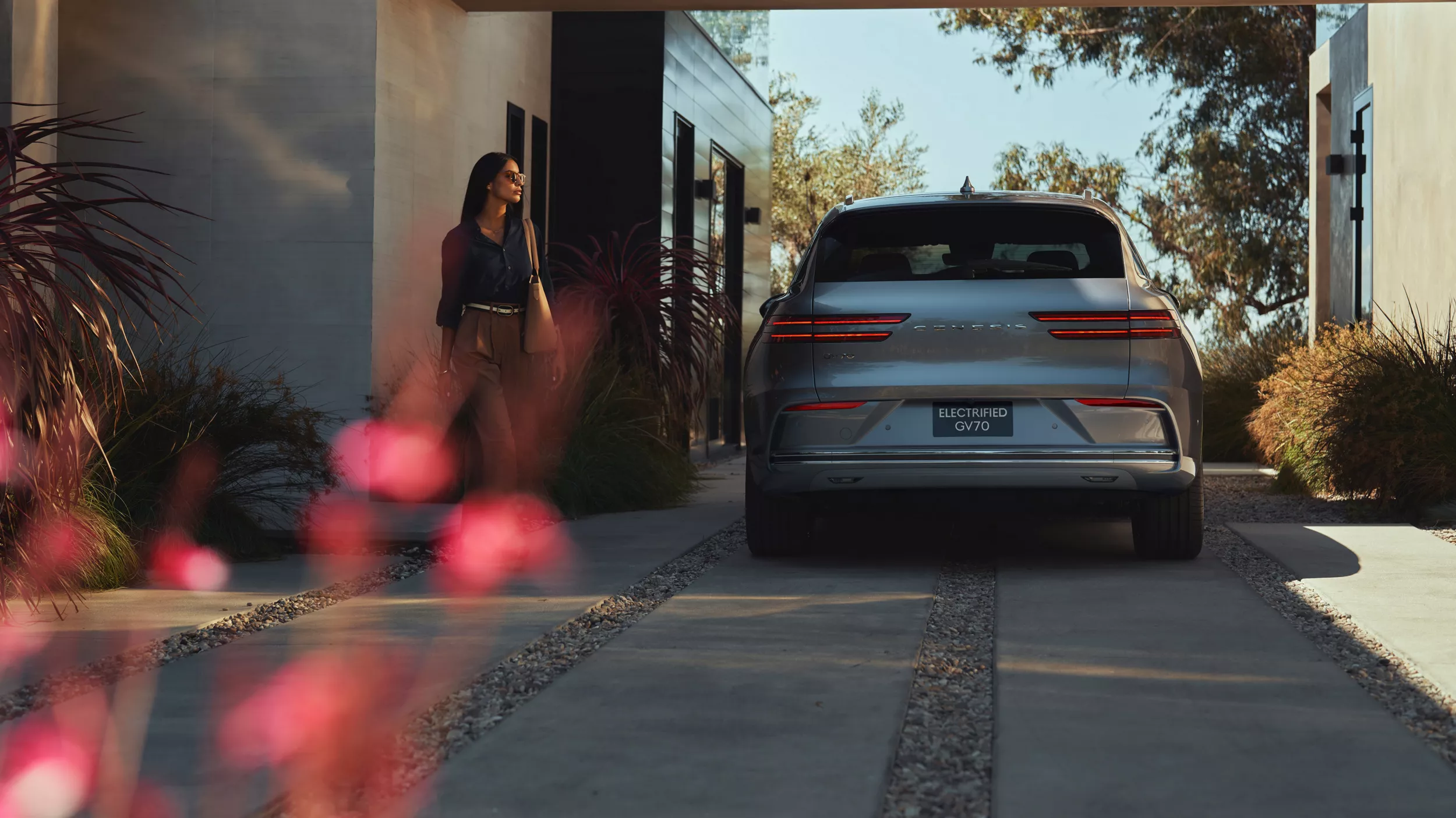 Woman walks toward driver side of Genesis Electrified GV70 in Savile Silver, parked in carport