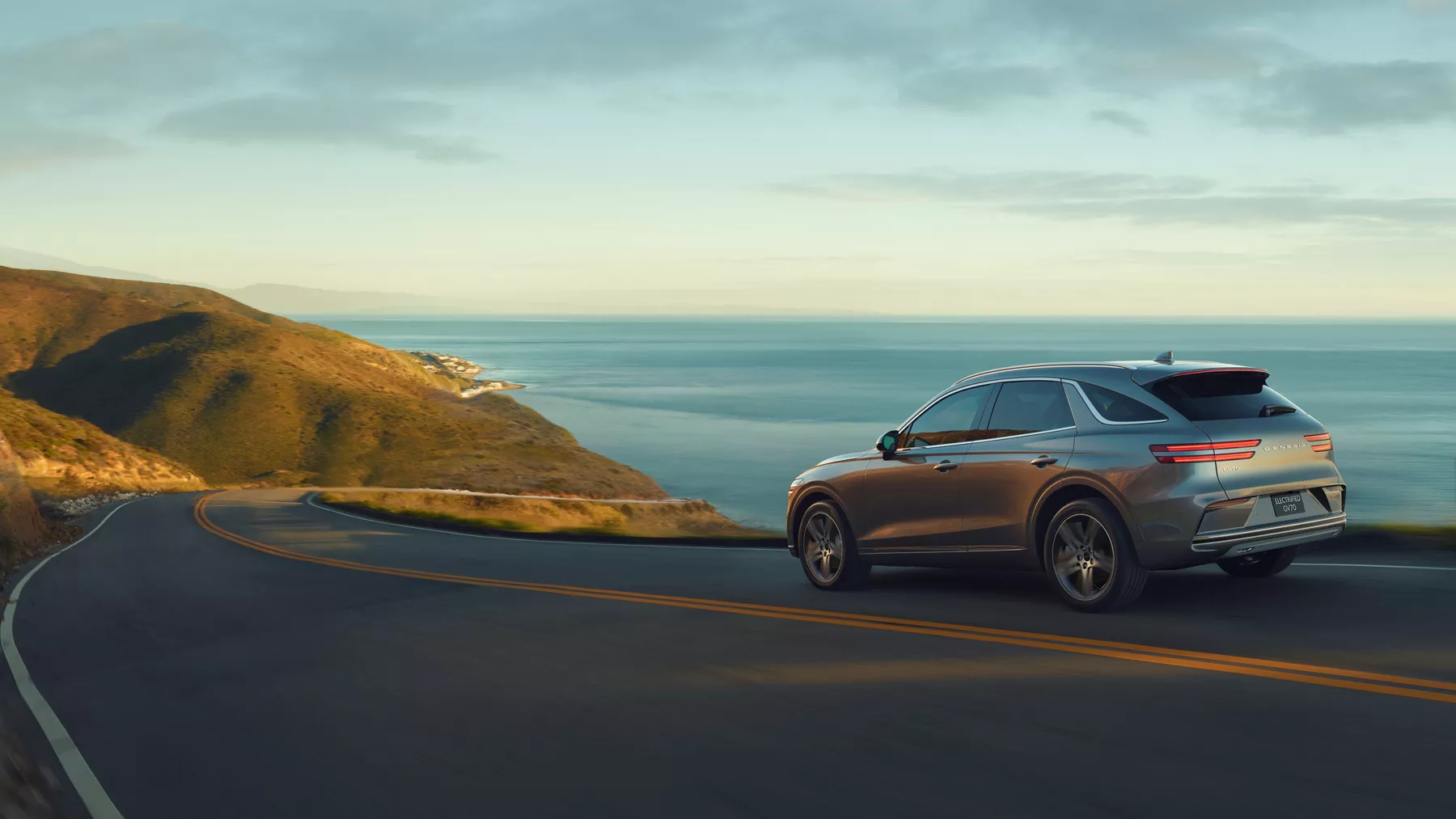 Rear driver side shot of 2026 Genesis Electrified GV70 in Savile Silver driving on a coastal highway