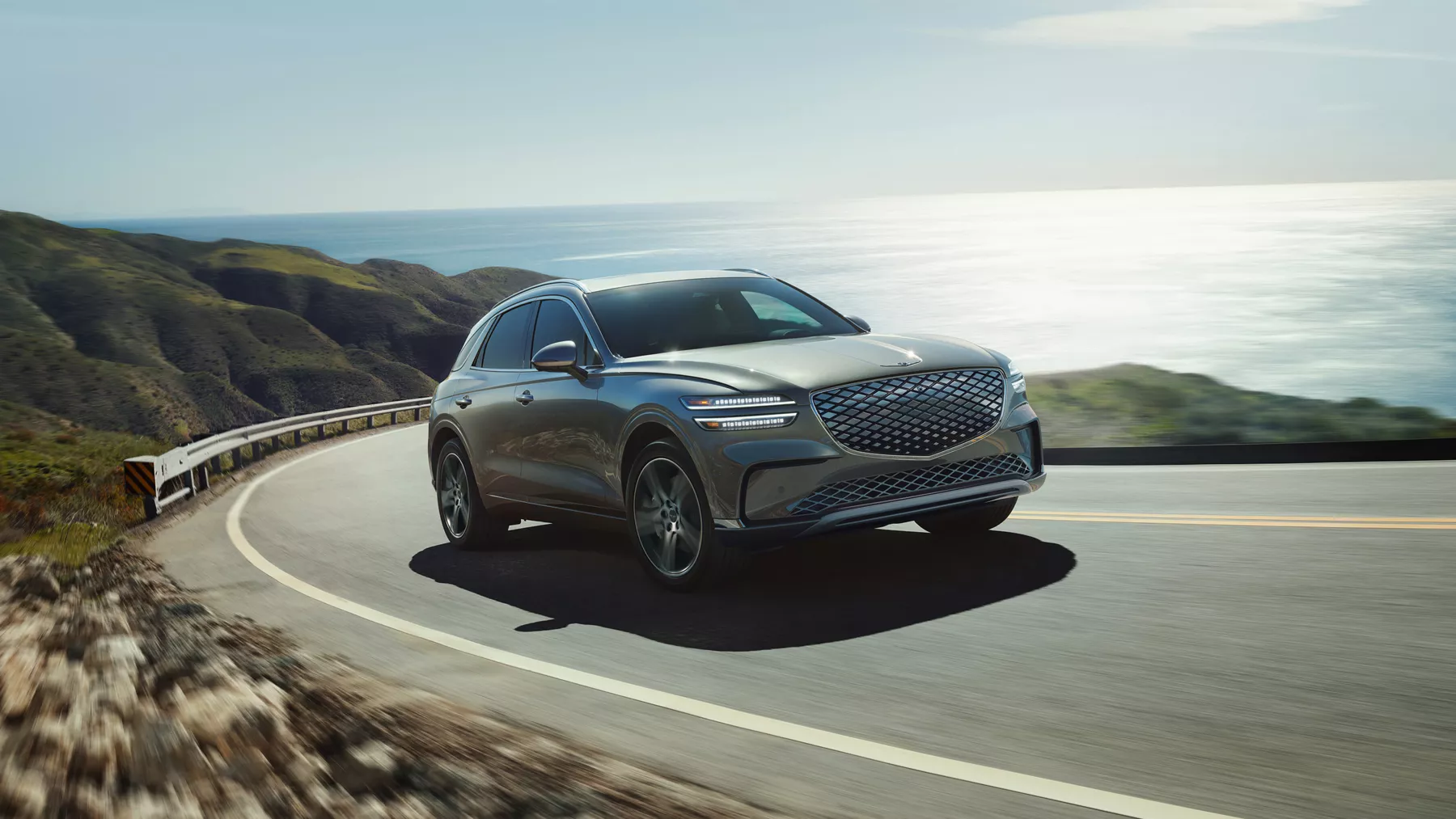 Front passenger side view of Genesis Electrified GV70 in Ceres Blue driving along coastal highway