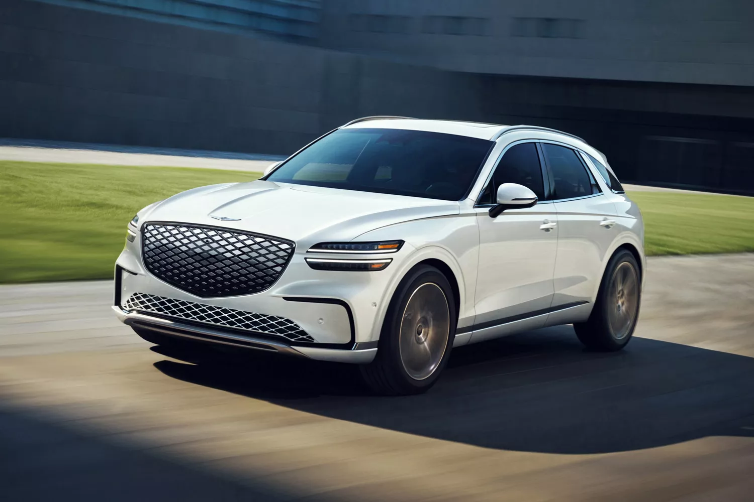 3/4 front driver side view Genesis Electrified GV70 in Uyuni White driving outside modern building