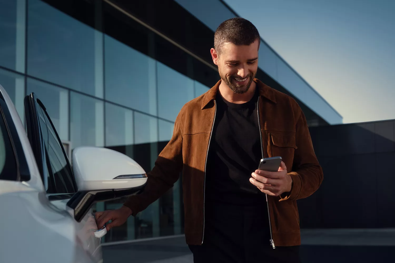 Man standing next to white Genesis vehicle, checking phone, grasping the driver side door handle