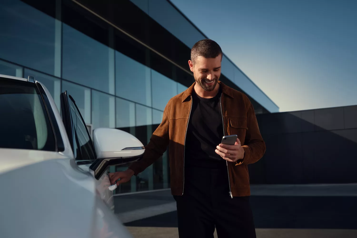 Person standing beside white 2026 Genesis GV70, holding a smartphone and opening the driver’s door