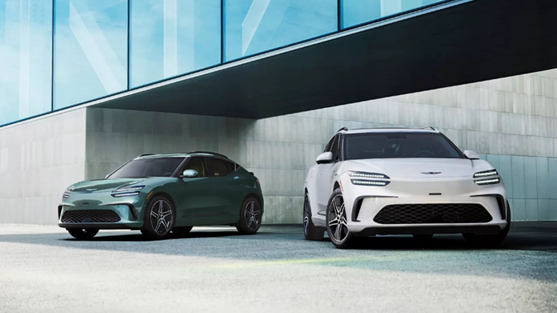 Two 2026 Genesis GV60 EVs in Tromso Green and Alta white parked under modern glass structure