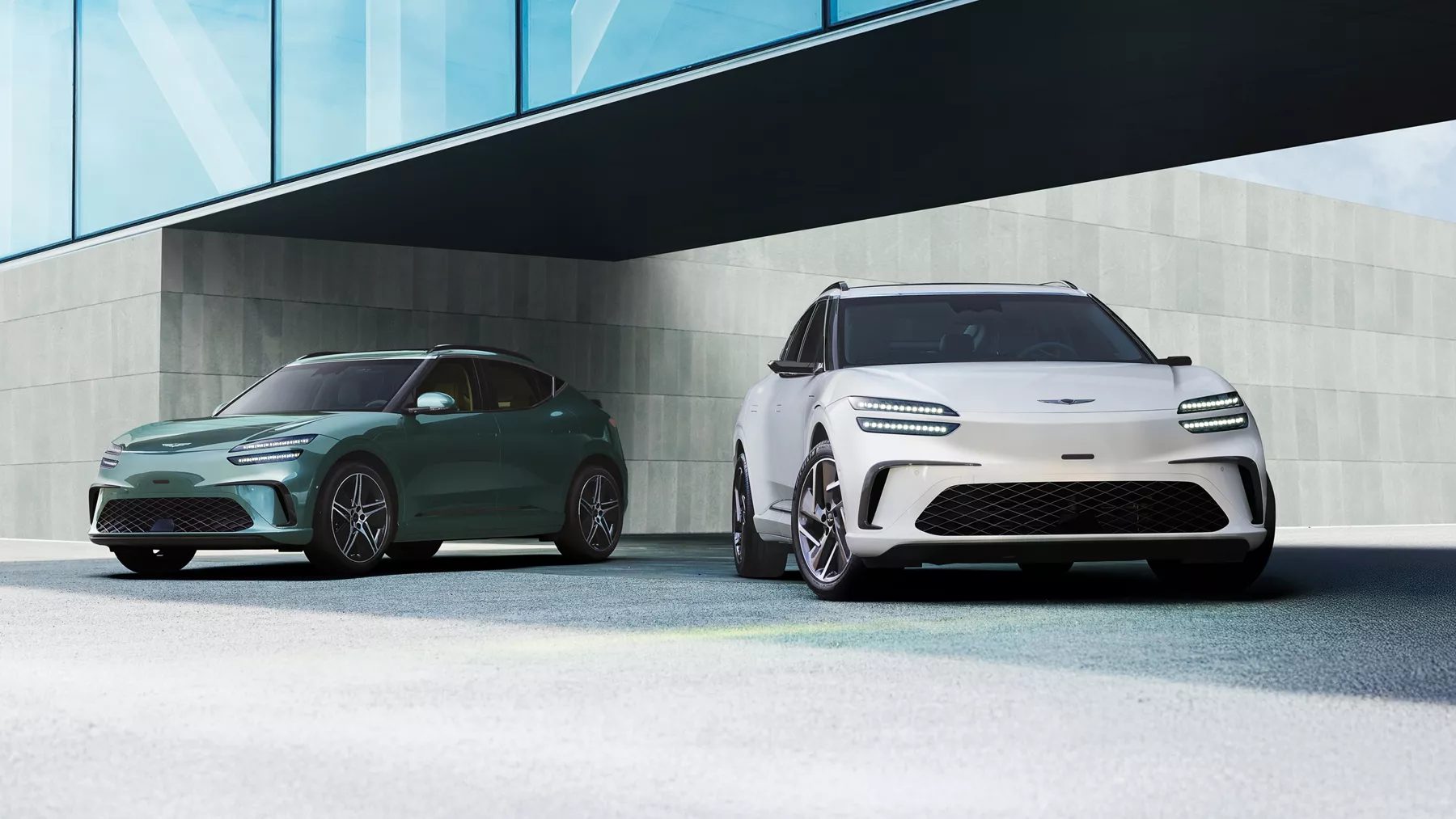 Two 2026 Genesis GV60 EVs in Tromso Green and Alta white parked under modern glass structure
