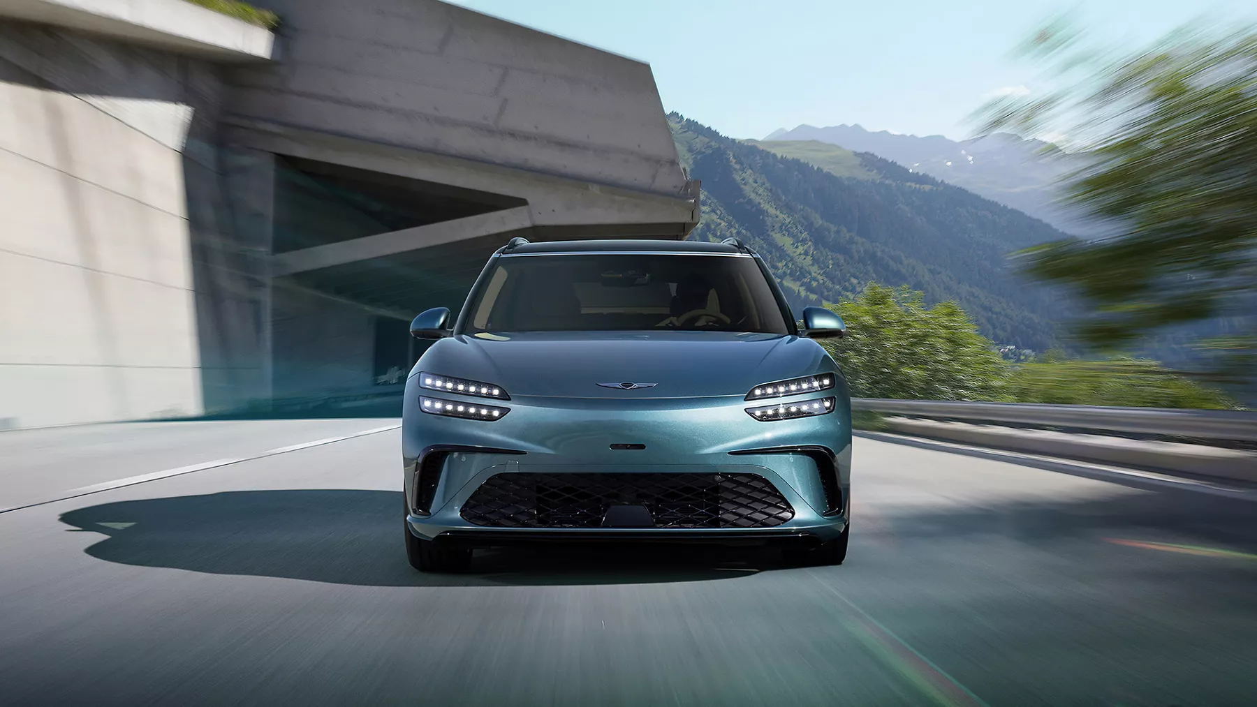 Front facing view of 2027 Genesis GV60 in Tromso Green driving mountainous road