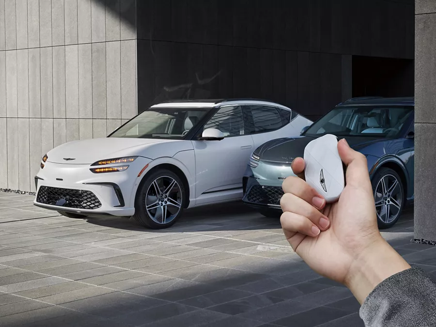 Two 2026 Genesis GV60's parked in modern garage, hand holding white Genesis key fob in foreground