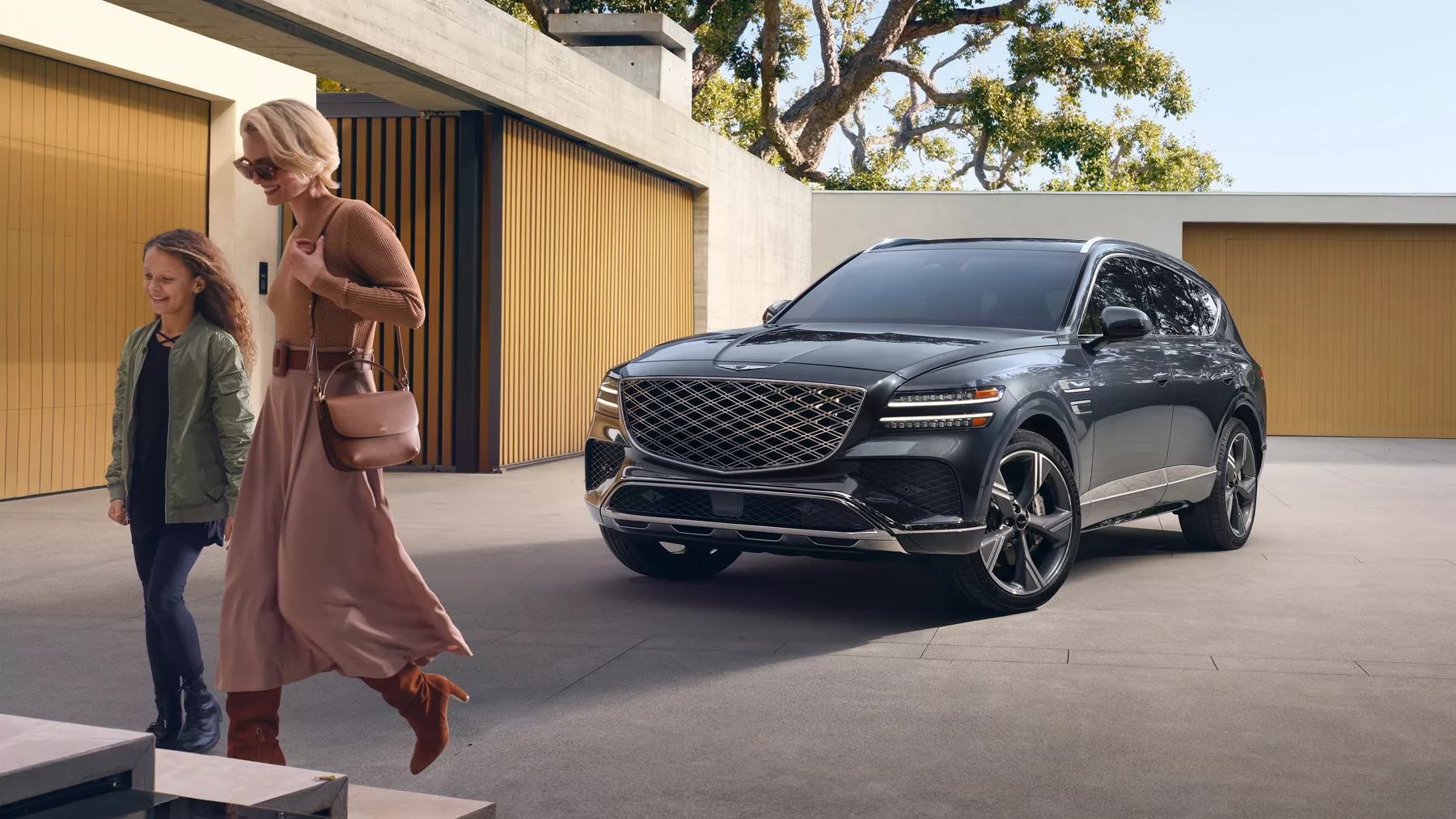 3/4 front driver side view of Makalu Gray Genesis GV80 beside modern home, mom & child walking past