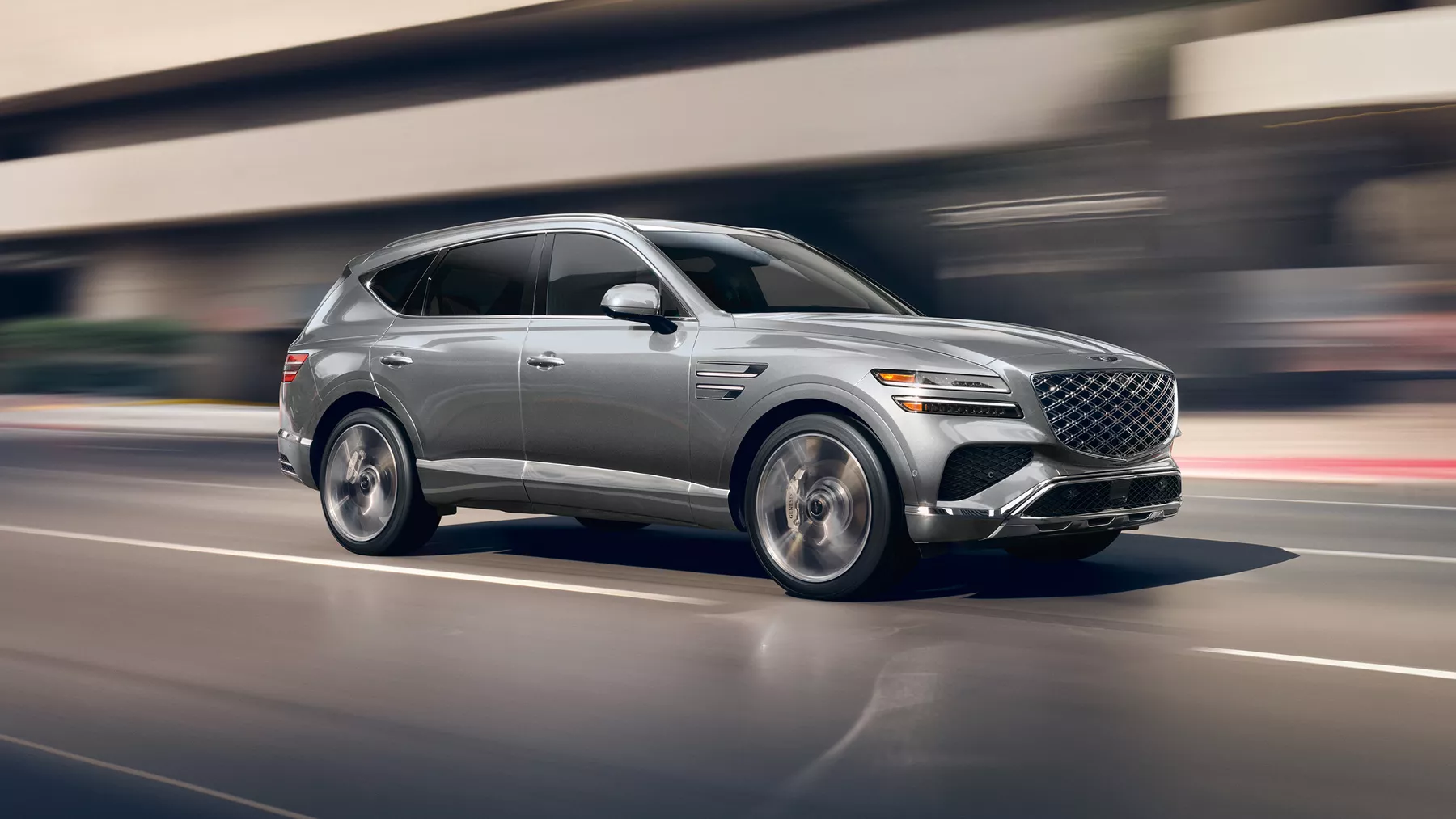 7/8 front passenger side view of motion blurred Genesis GV80 in Makalu Gray driving on city street