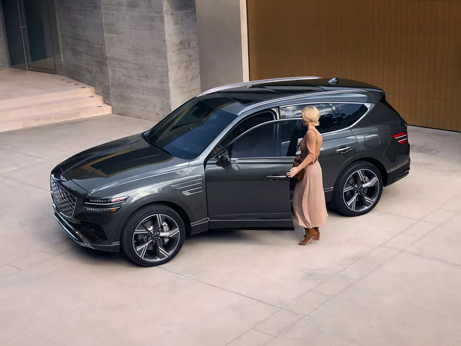 Aerial 3/4 driver side view of woman entering Genesis GV80 in Makalu Gray, parked at modern house