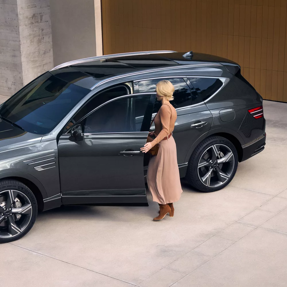 Aerial 3/4 driver side view of woman entering Genesis GV80 in Makalu Gray, parked at modern house