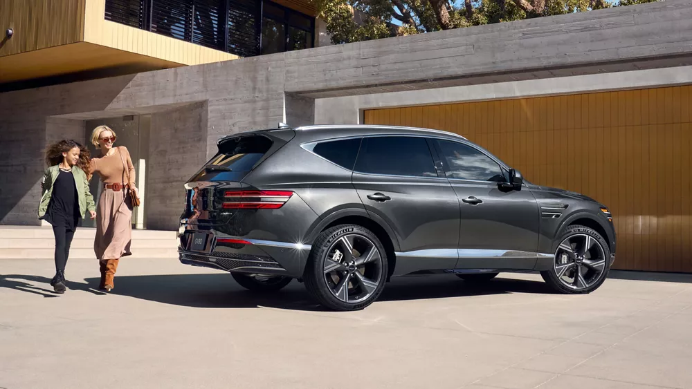 Passenger side profile view of woman and child walking behind Genesis GV80, parked at modern home