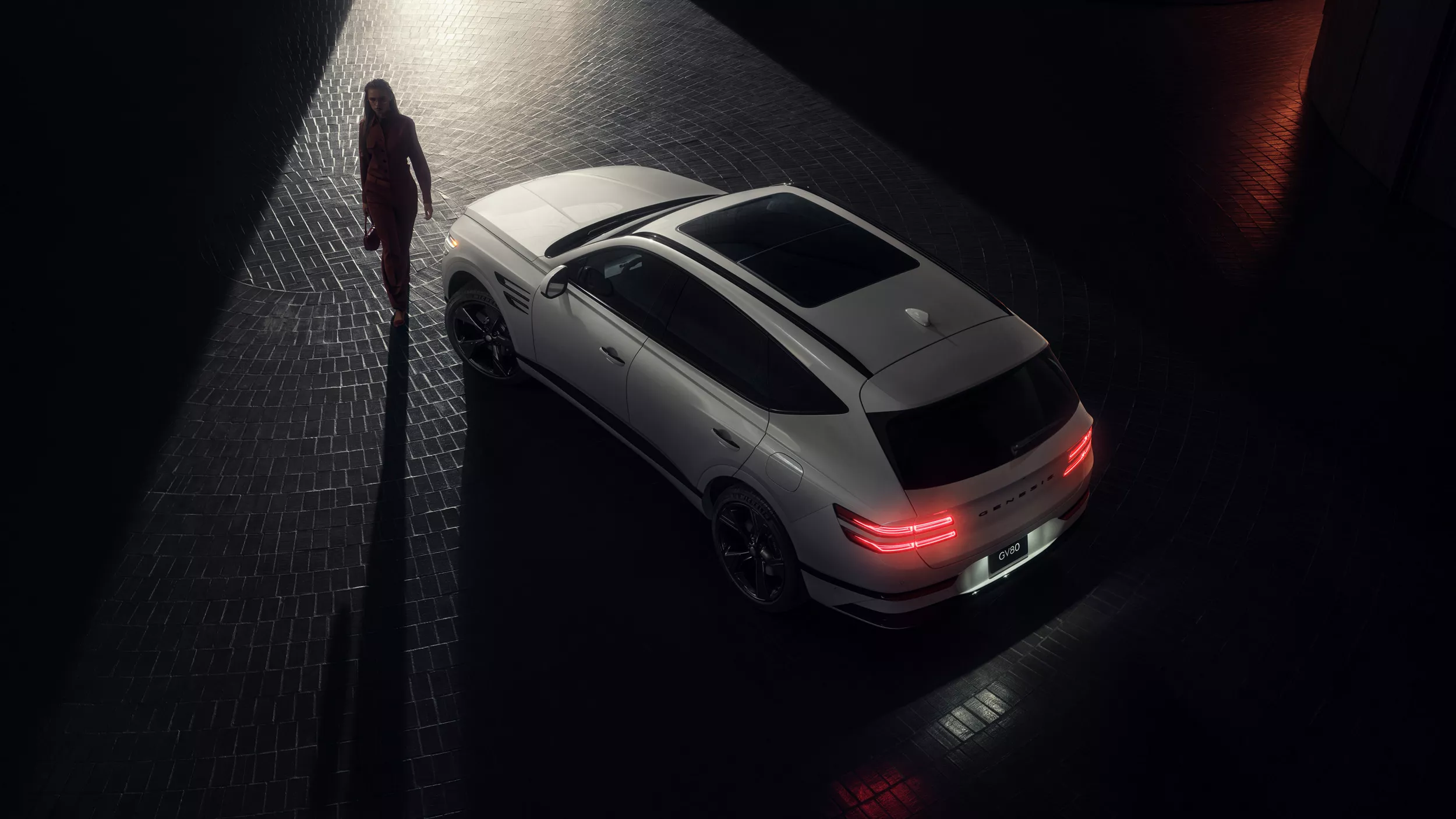 Aerial 3/4 rear driver side view of 2027 Genesis GV80 in dark space with person walking beside