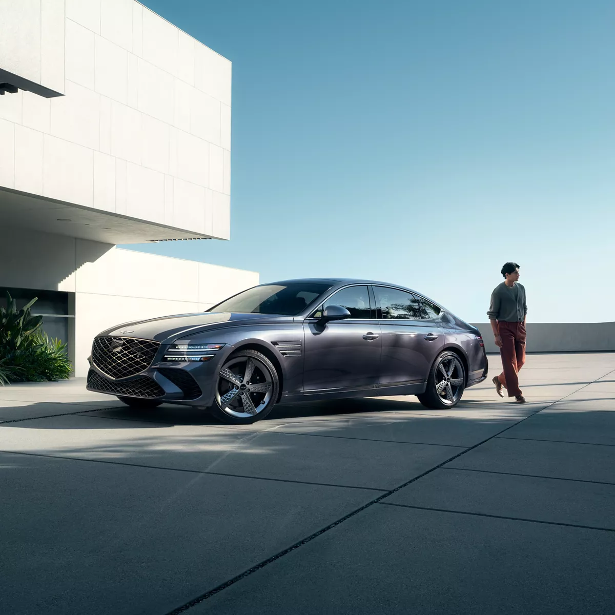 Front driver side view of 2026 Genesis G80 in Ceres Blue parked outside modern home as man walks by