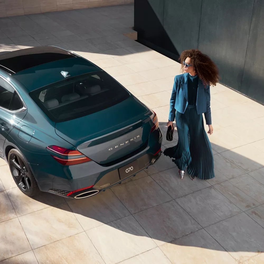 Aerial 3/4 rear driver side view of woman standing next to 2027 Genesis G70 in Tasman Blue