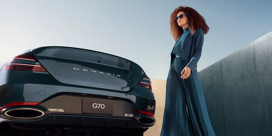 7/8 rear driver side view of 2027 Genesis G70 in Tasman Blue, woman in blue dress standing beside