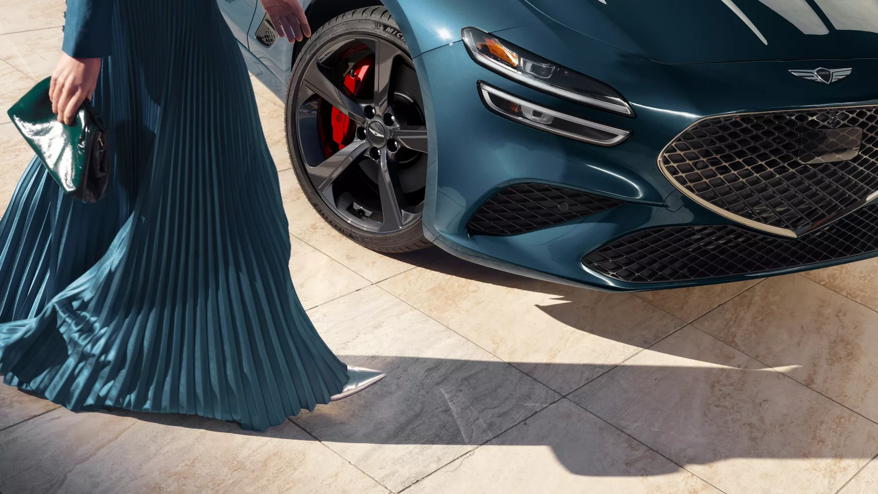 Close-up of wheel, headlight and grille on 2027 Genesis G70 in Tasman Blue, woman in dress nearby