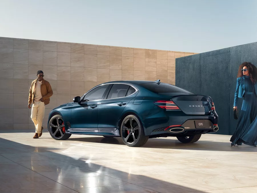 3/4 rear driver side view of 2027 Genesis G70 in Tasman Blue, man and woman standing on either side
