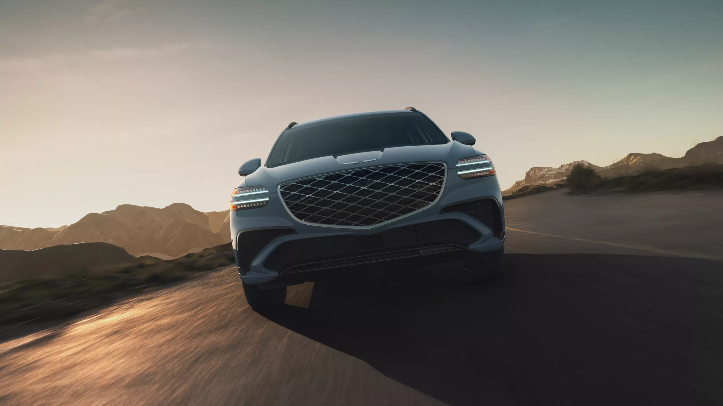 Exterior front ground level pov of Genesis GV70 driving on desert road with mountains in background