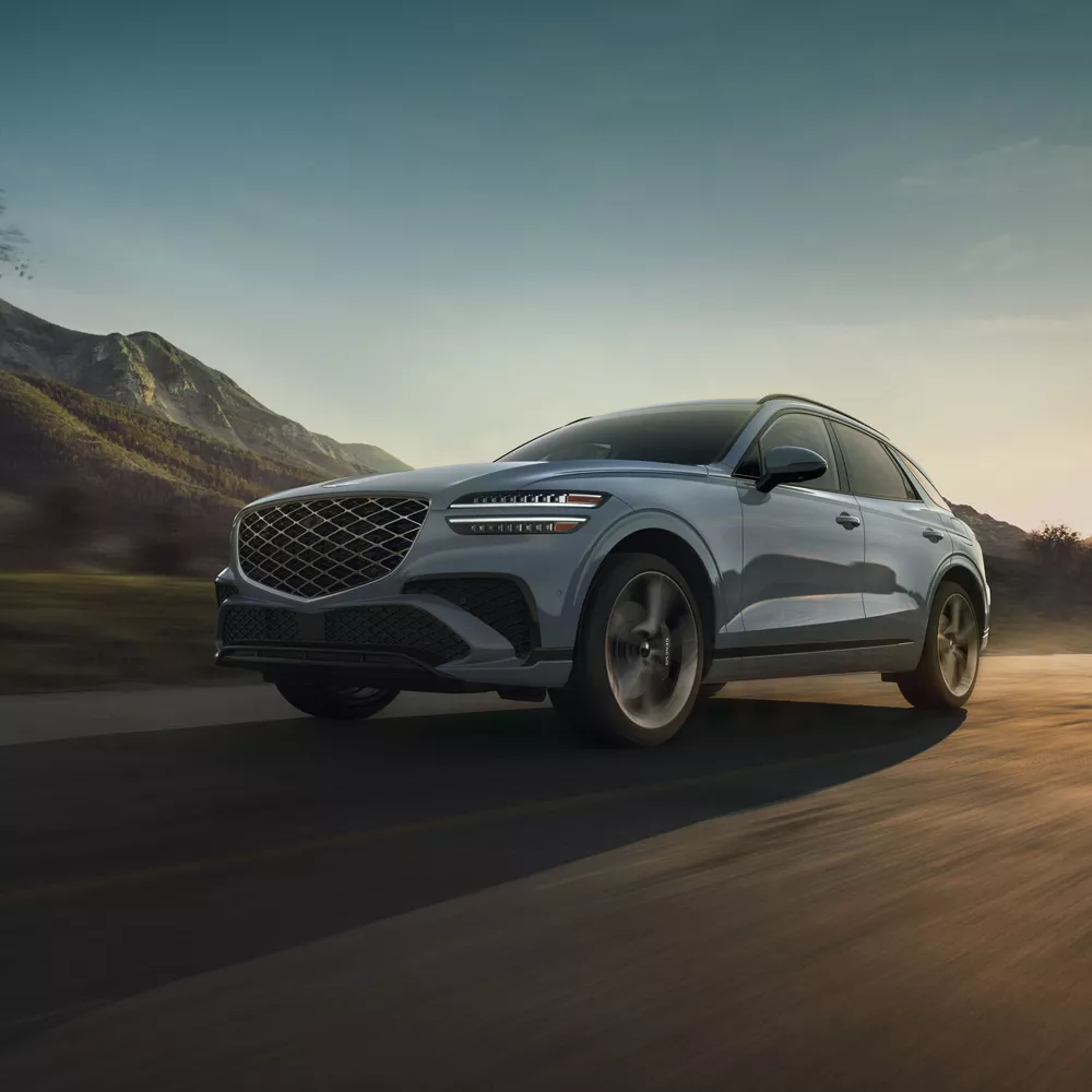 3/4 front driver side view of Genesis GV70 in Makalu Gray driving on desert road, mountains in back