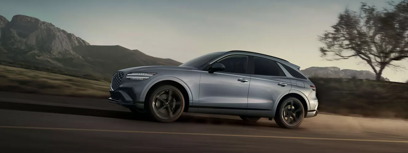 Driver side profile view of Genesis GV70 in Bering Blue road at dusk, with mountains in background