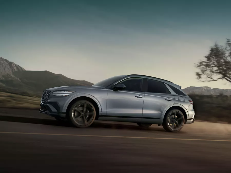 Driver side profile view of Genesis GV70 in Bering Blue road at dusk, with mountains in background