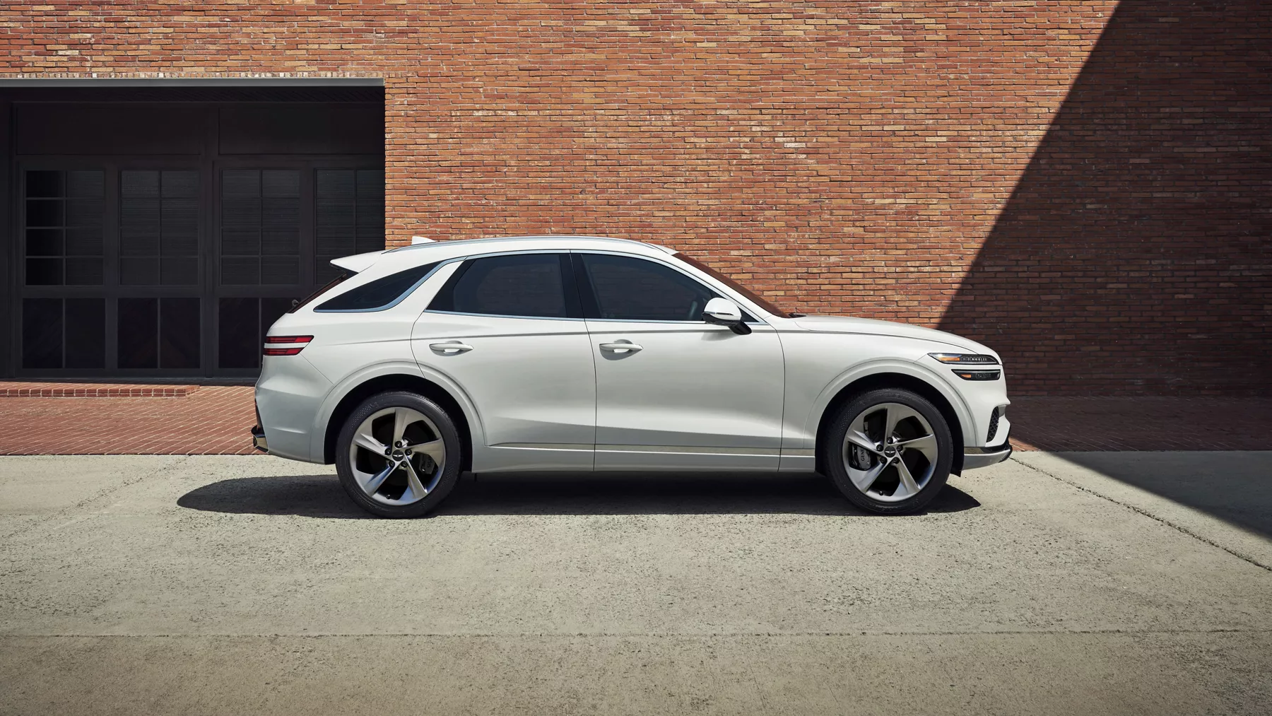 Passenger side profile view of Genesis GV70 in Uyuni White, parked beside brick building