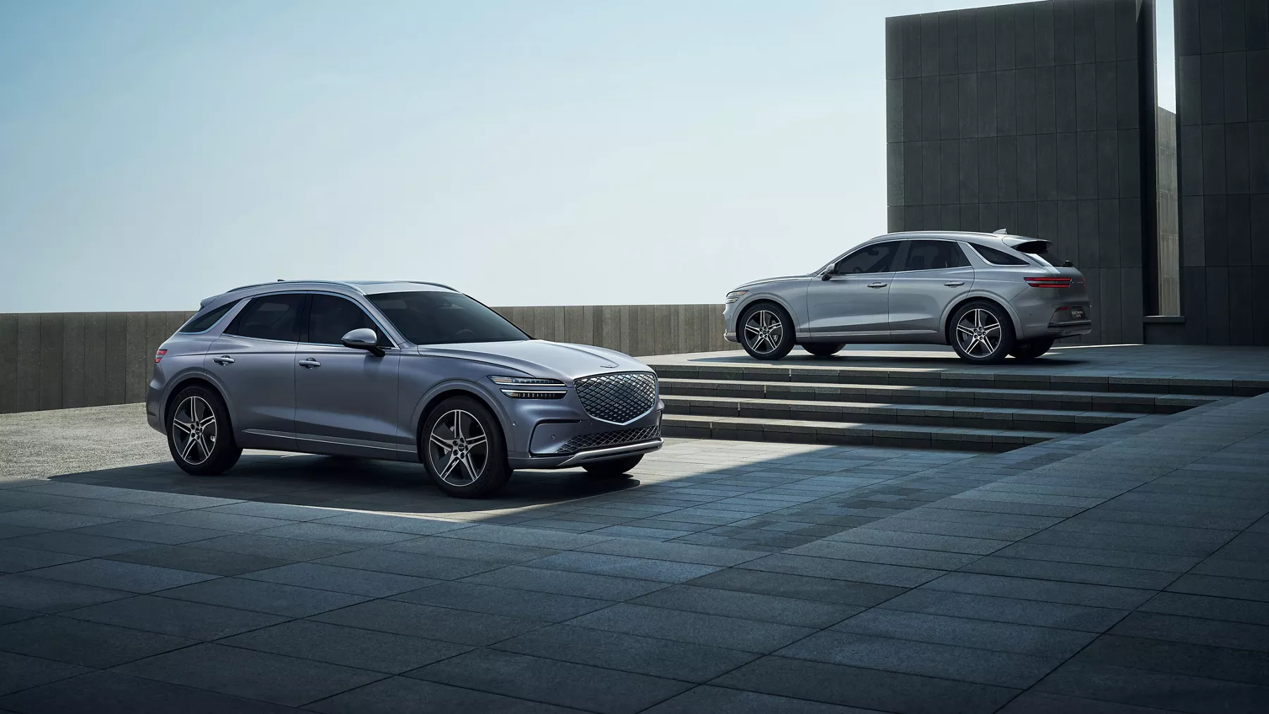 Two Genesis Electrified GV70 SUVs in Ceres Blue and Savile Silver parked in modern plaza