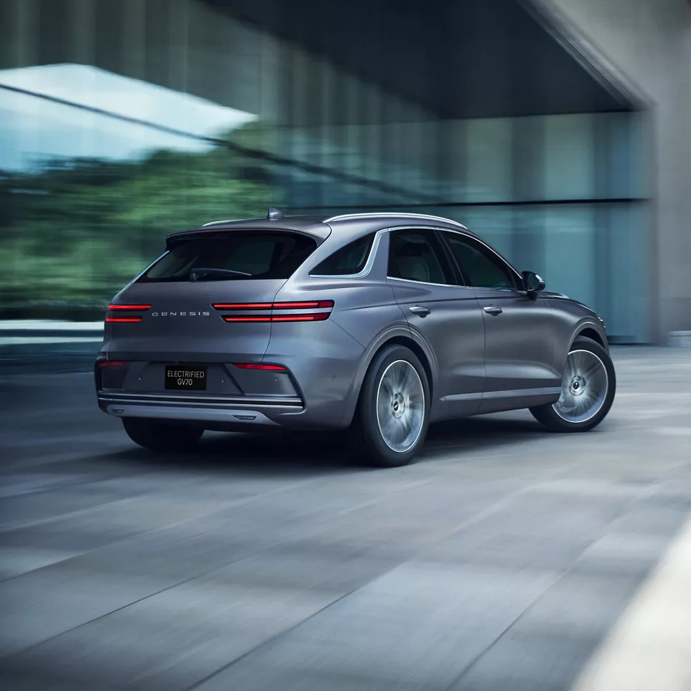 3/4 rear passenger side view of Ceres Blue Genesis Electrified GV70 in motion beside modern building