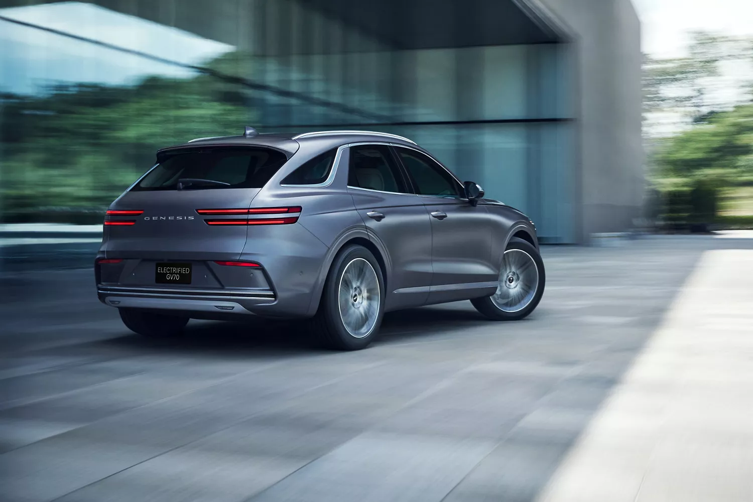 3/4 rear passenger side view of Ceres Blue Genesis Electrified GV70 in motion beside modern building