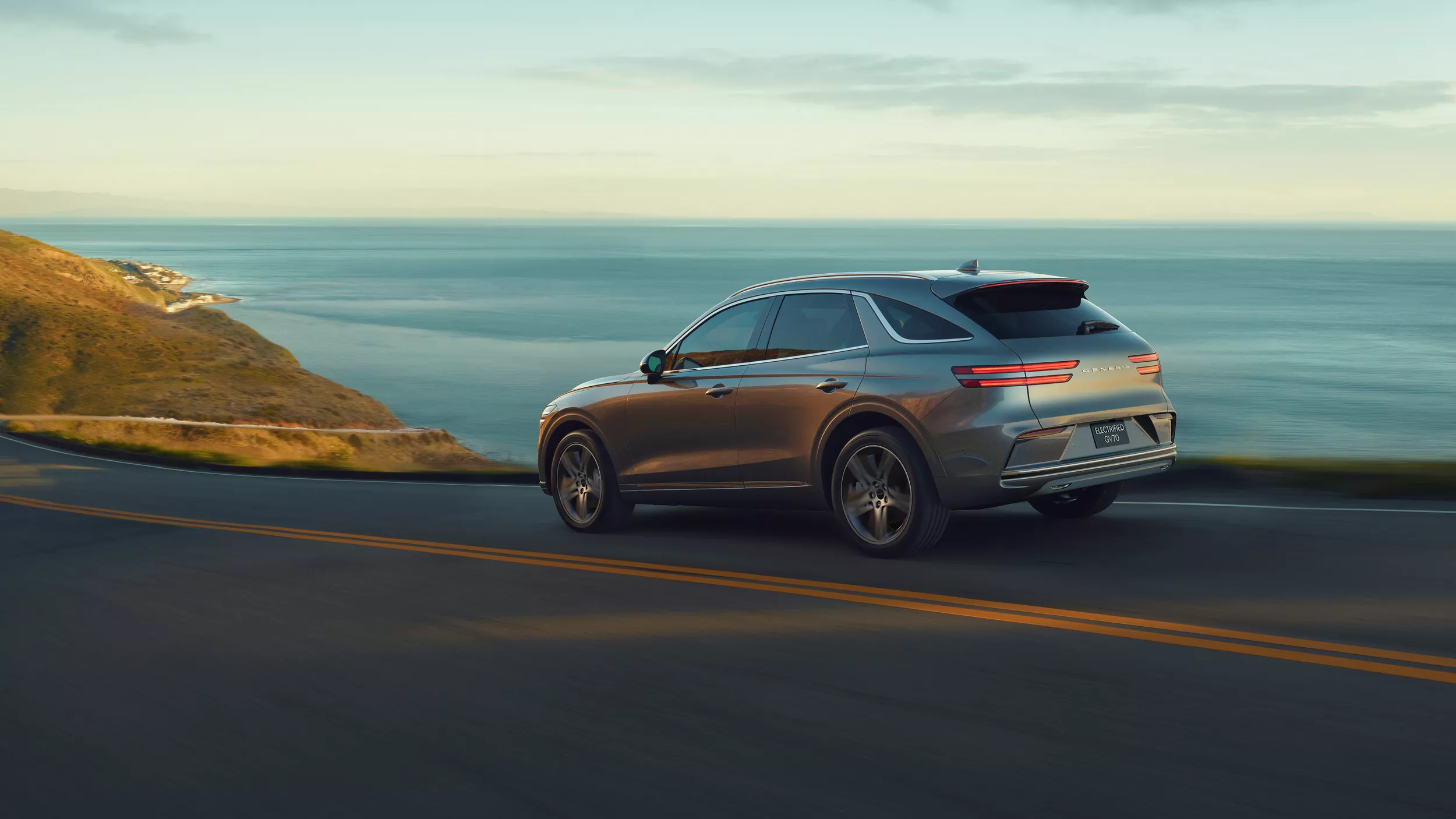 7/8 Rear Driver Side Genesis Electrified GV70 Prestige in Savile Silver in motion on coastal highway
