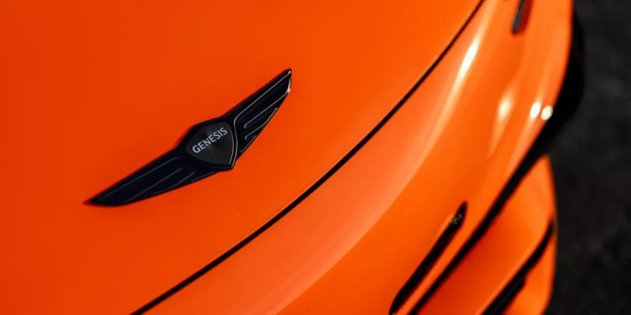 Closeup of black Genesis badge emblem on hood of 2027 Genesis GV60 Magma in orange