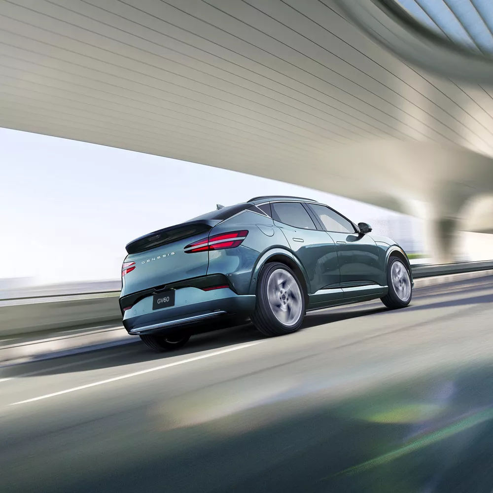 3/4 rear passenger side view of Genesis GV60 in Tromso Green, diving under futuristic bridge in city