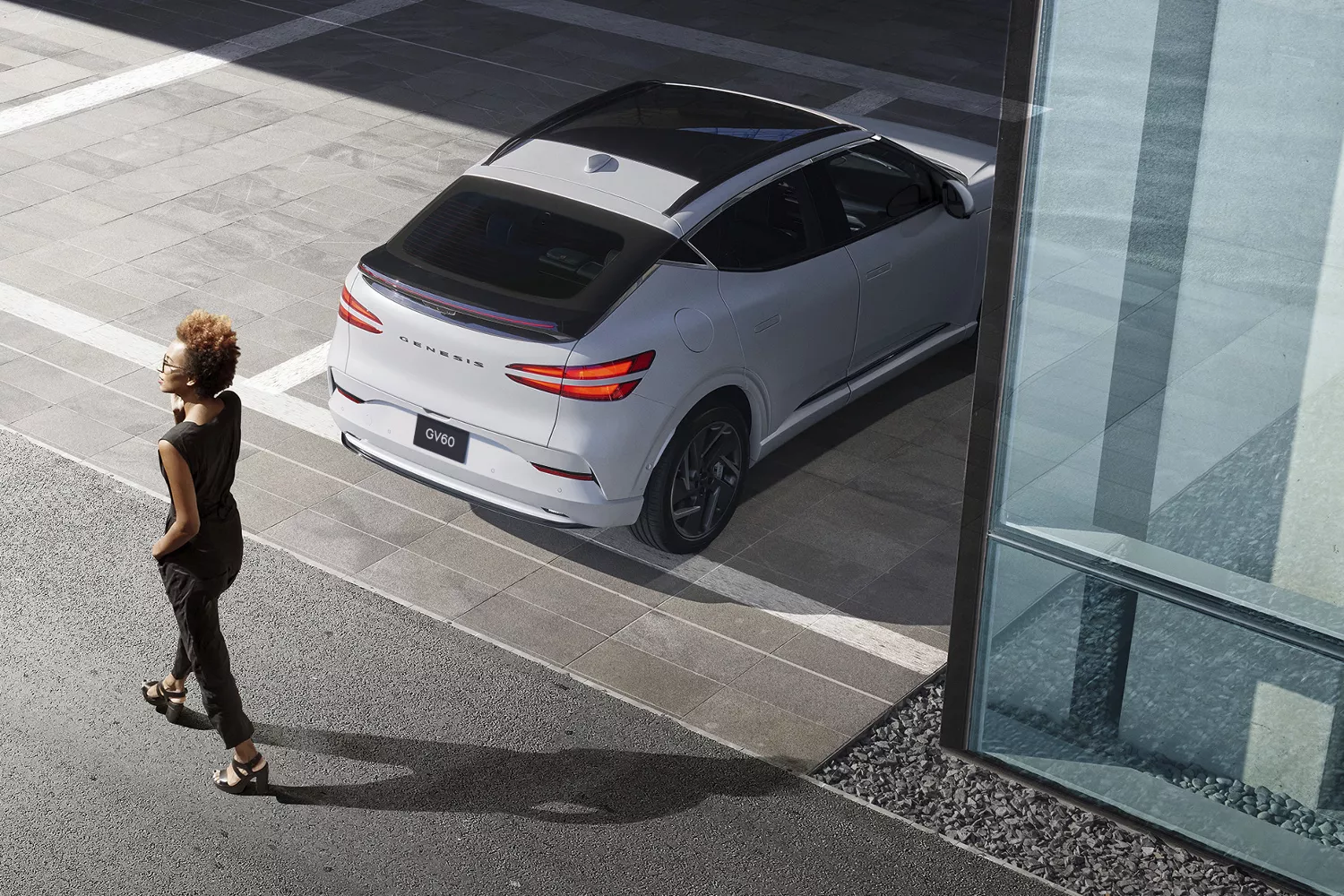 3/4 rear aerial view of woman walking past Genesis GV60 in Uyuni White parked in modern carpor